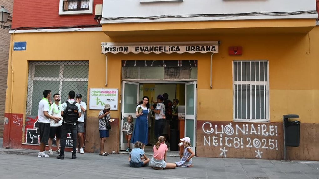 Vor einem gelben Gebäude mit der Aufschrift „Misión Evangélica Urbana“ stehen und sitzen Menschen. Kinder sitzen auf dem Bürgersteig.