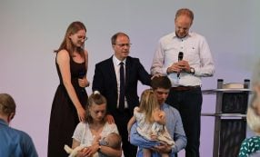 Fünf Erwachsene und zwei Kinder nehmen an einer kirchlichen Feier teil; zwei Männer halten Mikrofone in der Hand und sprechen ein Gebet für eine Familie.
