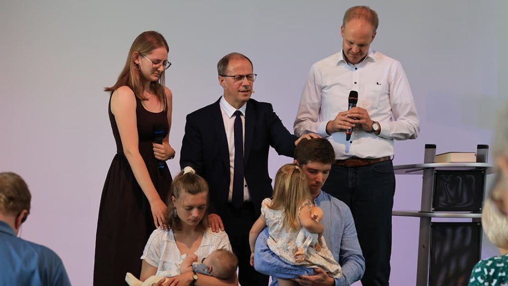 Fünf Erwachsene und zwei Kinder nehmen an einer kirchlichen Feier teil; zwei Männer halten Mikrofone in der Hand und sprechen ein Gebet für eine Familie.
