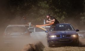 Zwei Männer duellieren sich mit Schwimmnudeln, während sie auf fahrenden Autos in einer staubigen Umgebung im Freien stehen.