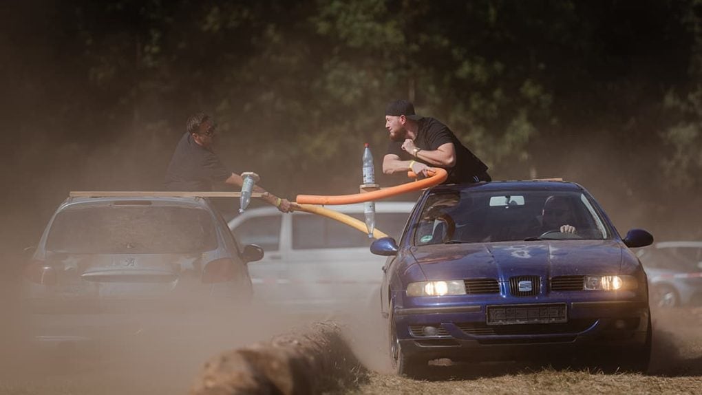 Zwei Männer duellieren sich mit Schwimmnudeln, während sie auf fahrenden Autos in einer staubigen Umgebung im Freien stehen.
