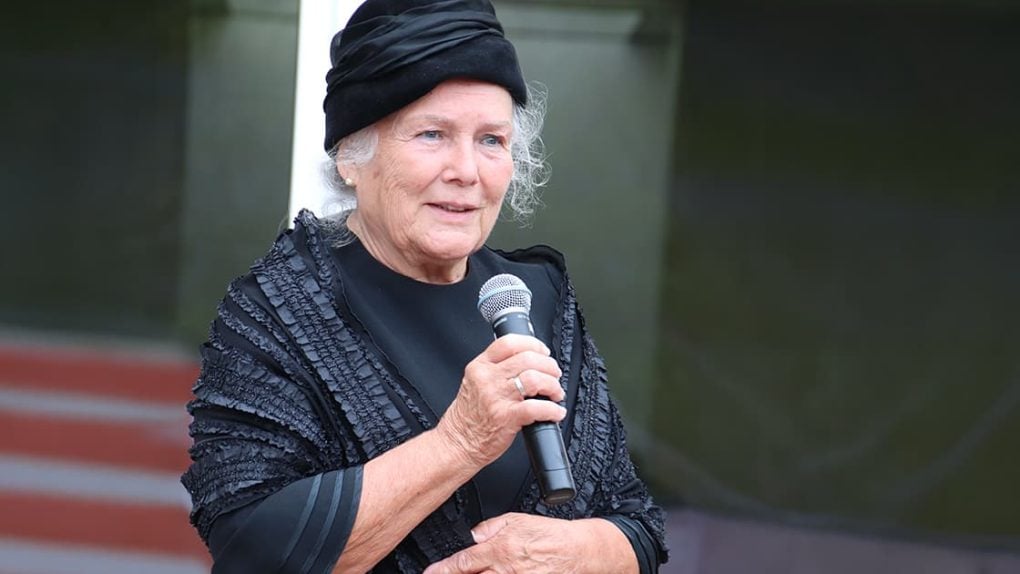 Judi Dench trägt einen schwarzen Hut und einen Schal und hält ein Mikrofon in der Hand, während sie spricht.