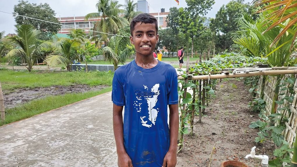 Ein lächelnder Junge in einem blauen T-Shirt steht auf einem Weg in einem Garten.