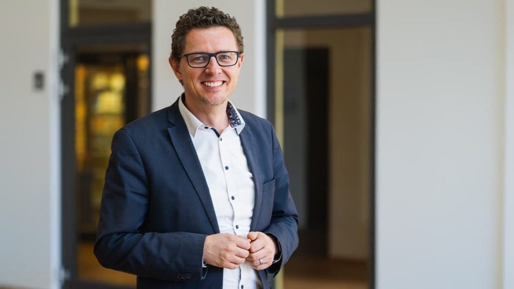 Ein lächelnder Mann in Blazer und Hemd, der in einem Büroflur steht.