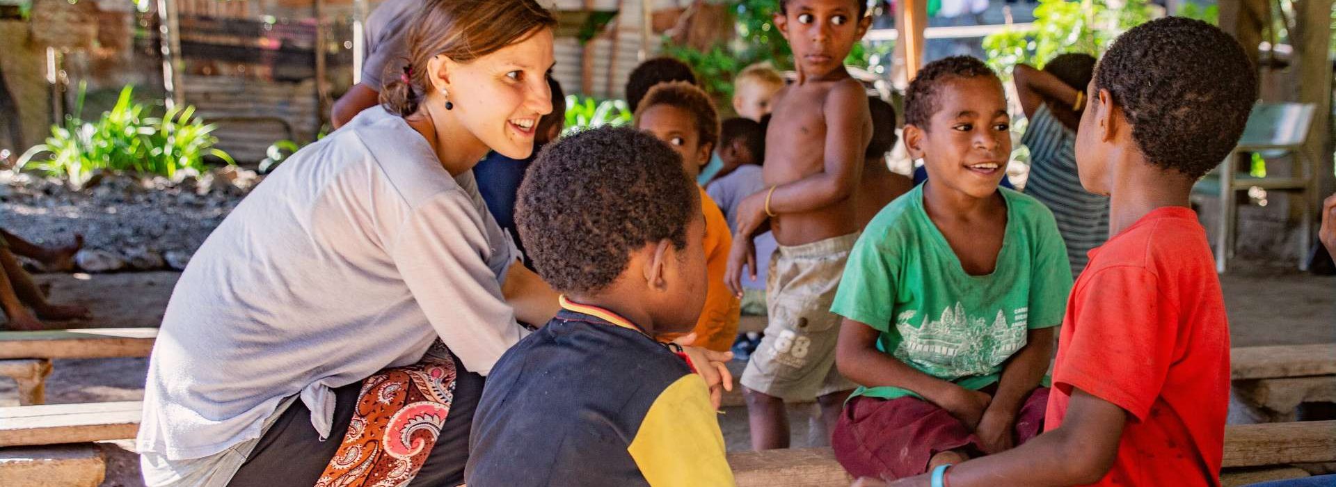 Eine Frau sitzt da und lächelt, während sie sich im Freien in einer tropischen, ländlichen Umgebung mit mehreren Kindern unterhält.