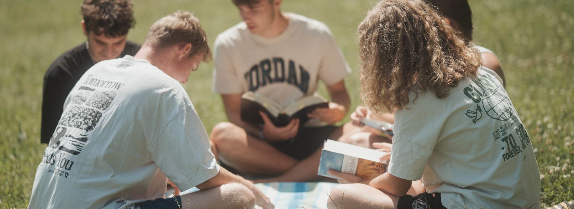 Fünf Menschen sitzen auf dem Rasen und lesen gemeinsam auf einer gestreiften Decke im Sonnenlicht Bücher.