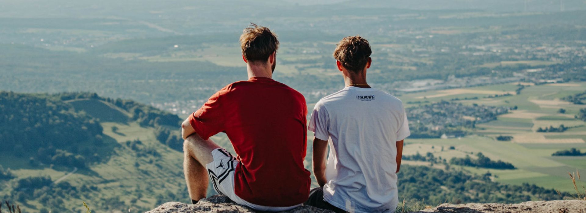 An einem klaren Tag sitzen zwei Menschen auf einem Felsvorsprung und blicken auf eine malerische Landschaft aus Feldern und Hügeln.