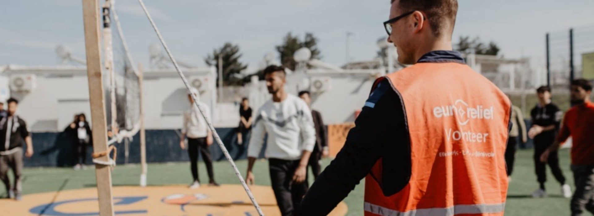 Ein Freiwilliger in einer orangefarbenen Weste beobachtet, wie Menschen auf einem Außenplatz Volleyball spielen.