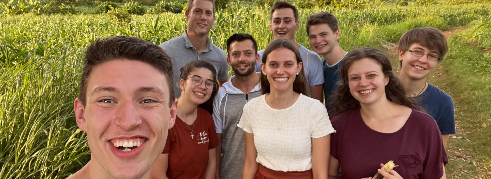 Neun lächelnde Menschen stehen gemeinsam draußen auf einem grasbewachsenen Weg, im Hintergrund sind Berge zu sehen.