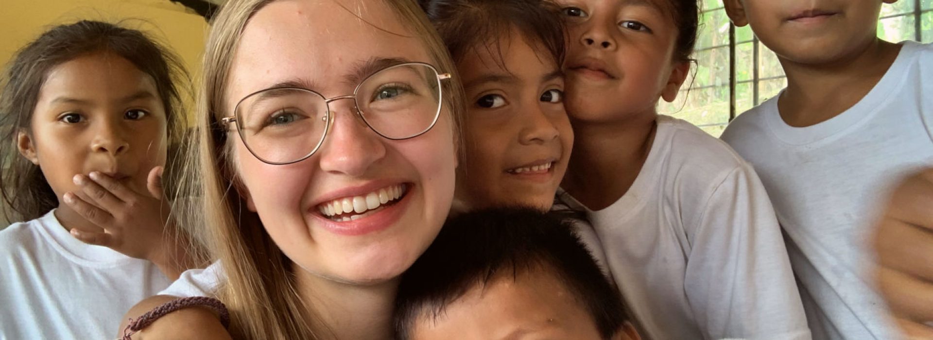 Eine lächelnde junge Frau mit Brille, umgeben von fünf Kindern in einem hellen Raum; alle blicken in die Kamera.