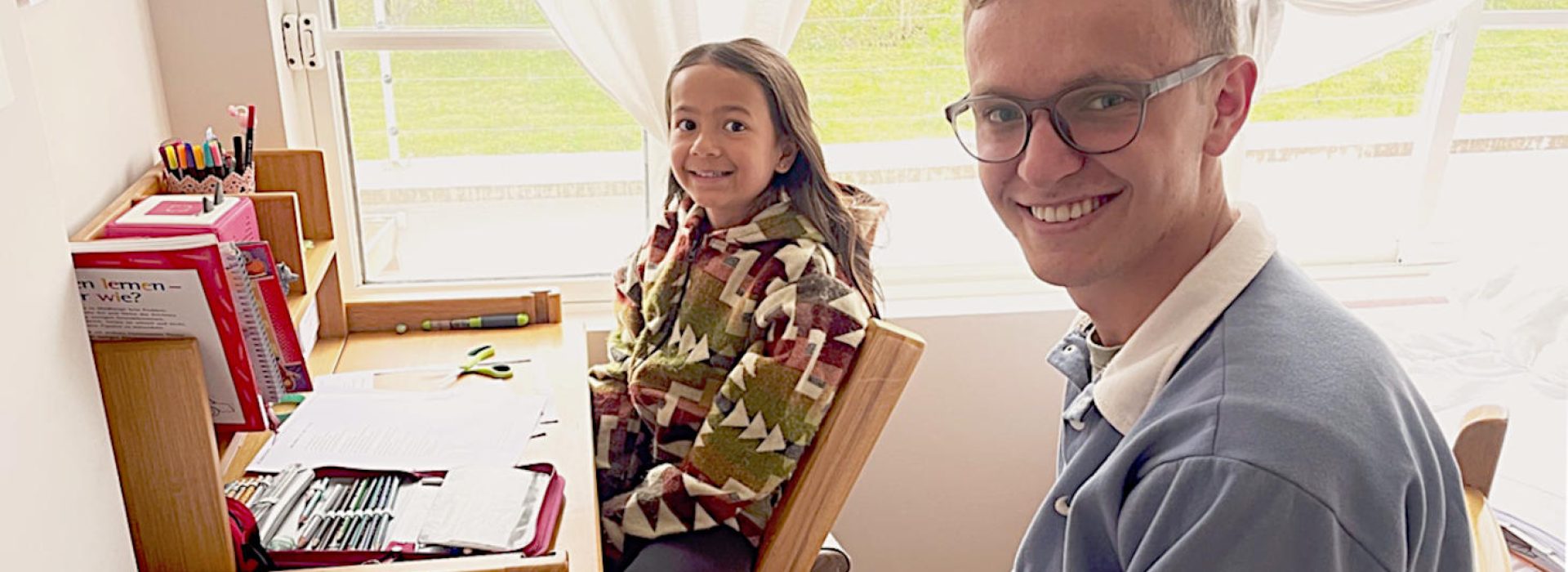 Ein lächelnder Mann und ein Mädchen sitzen an einem Schreibtisch mit Papieren und Schulmaterial in der Nähe eines hellen Fensters.