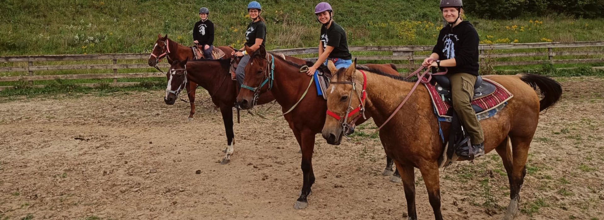 Vier Personen mit Helmen und schwarzen Hemden sitzen auf Pferden in einem eingezäunten Reitplatz im Freien, hinter ihnen liegen grasbewachsene Hügel.