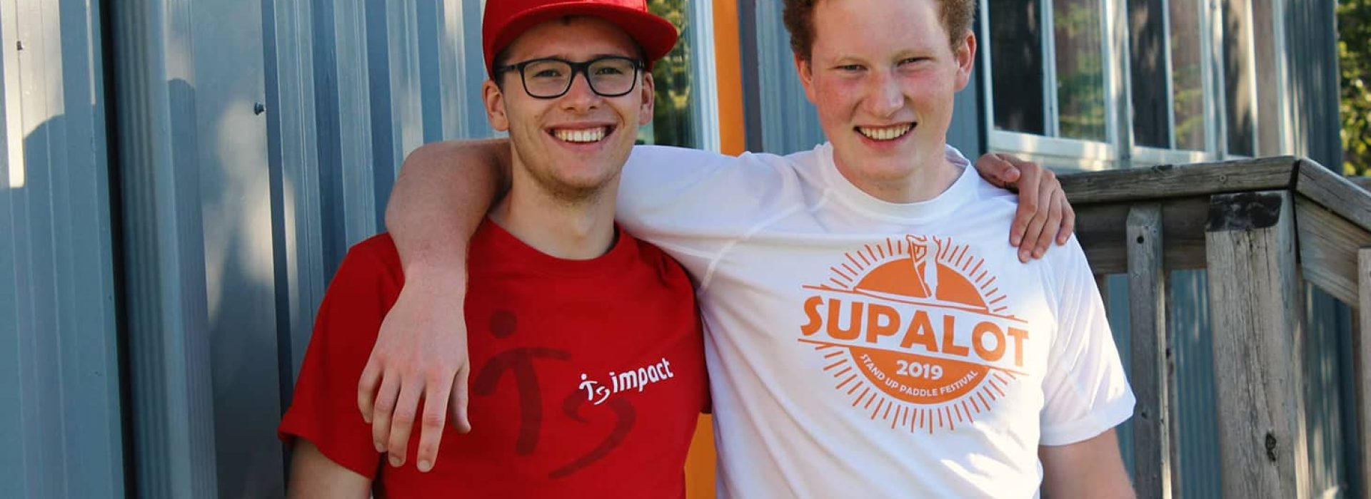 Zwei lächelnde junge Männer, einer in einem roten Hemd und einer mit einer roten Mütze, der andere in einem weißen Hemd, stehen Seite an Seite und haben ihre Arme umeinander gelegt.