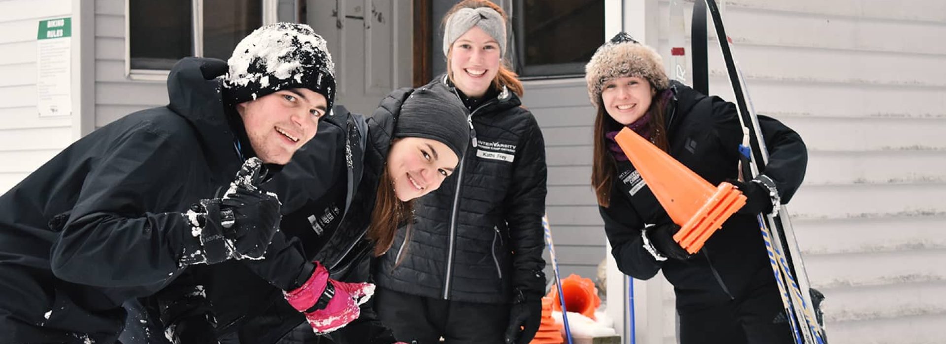 Vier Personen in Winterkleidung lächeln im Freien, halten Skistöcke und orangefarbene Kegel in den Händen, und auf ihren Mützen und Jacken liegt Schnee.