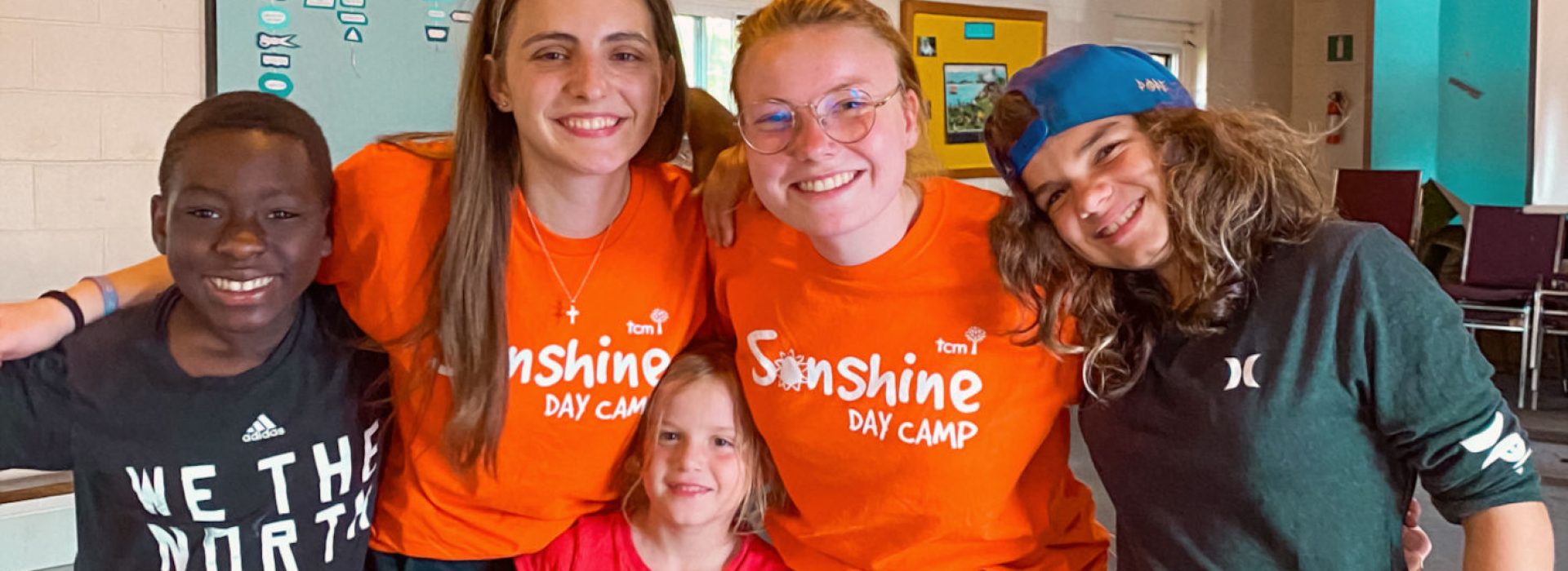 Fünf lächelnde Kinder und Jugendliche, von denen zwei orangefarbene „Sunshine Day Camp“-T-Shirts tragen, posieren gemeinsam in einem Raum und haben ihre Arme umeinander gelegt.