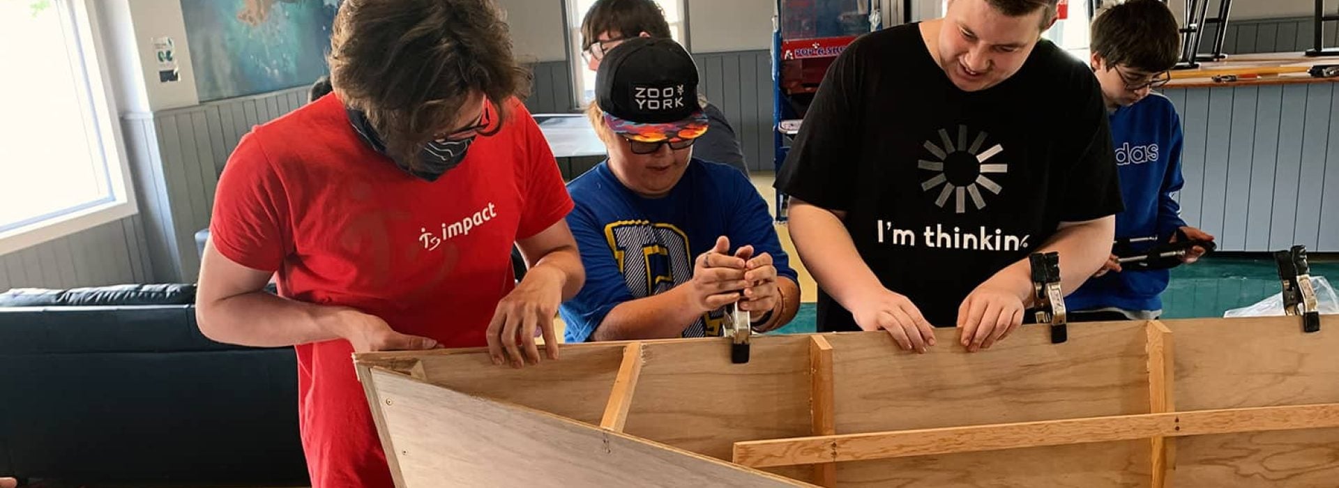 Drei Jugendliche bauen in einem Raum mit Hilfe von Klemmen und Werkzeugen eine Holzkonstruktion zusammen und sind ganz auf ihr praktisches Projekt konzentriert.