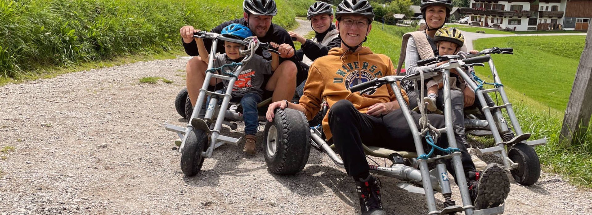 Fünf Personen mit Helmen sitzen auf Pedal-Go-Karts auf einem Schotterweg in einer malerischen, ländlichen Gegend mit Wiesen und Gebäuden.