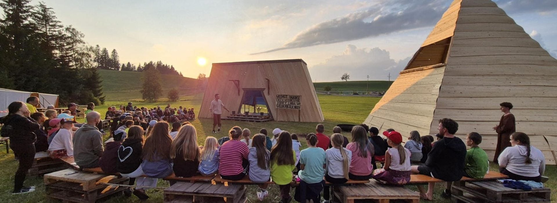 Kinder sitzen auf Bänken und beobachten bei Sonnenuntergang einen Künstler im Freien in der Nähe moderner Holzkonstruktionen.