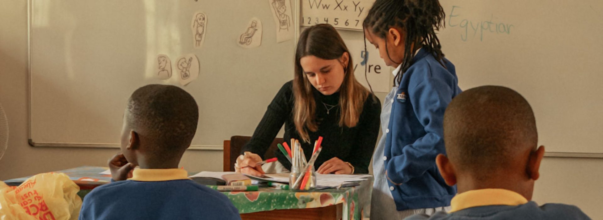 Eine Lehrerin sitzt an einem Tisch und hilft einem Schüler, während zwei andere Kinder in der Nähe in einem Klassenzimmer sitzen.