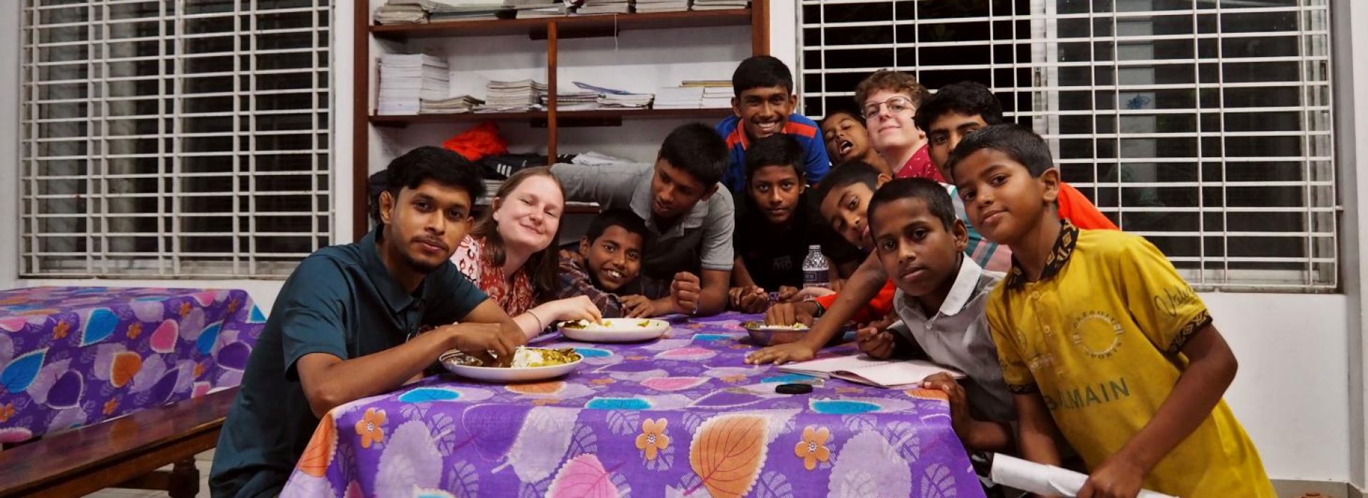 Eine Gruppe von Jugendlichen und ein Erwachsener sitzen um einen Tisch mit Essen herum und lächeln in einem hellen Raum in die Kamera.