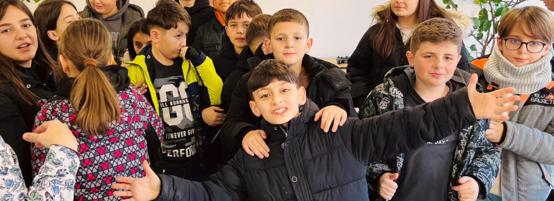 Eine Gruppe von Kindern steht lächelnd zusammen in einem Raum, wobei ein Junge ganz vorne steht und die Arme weit ausbreitet.