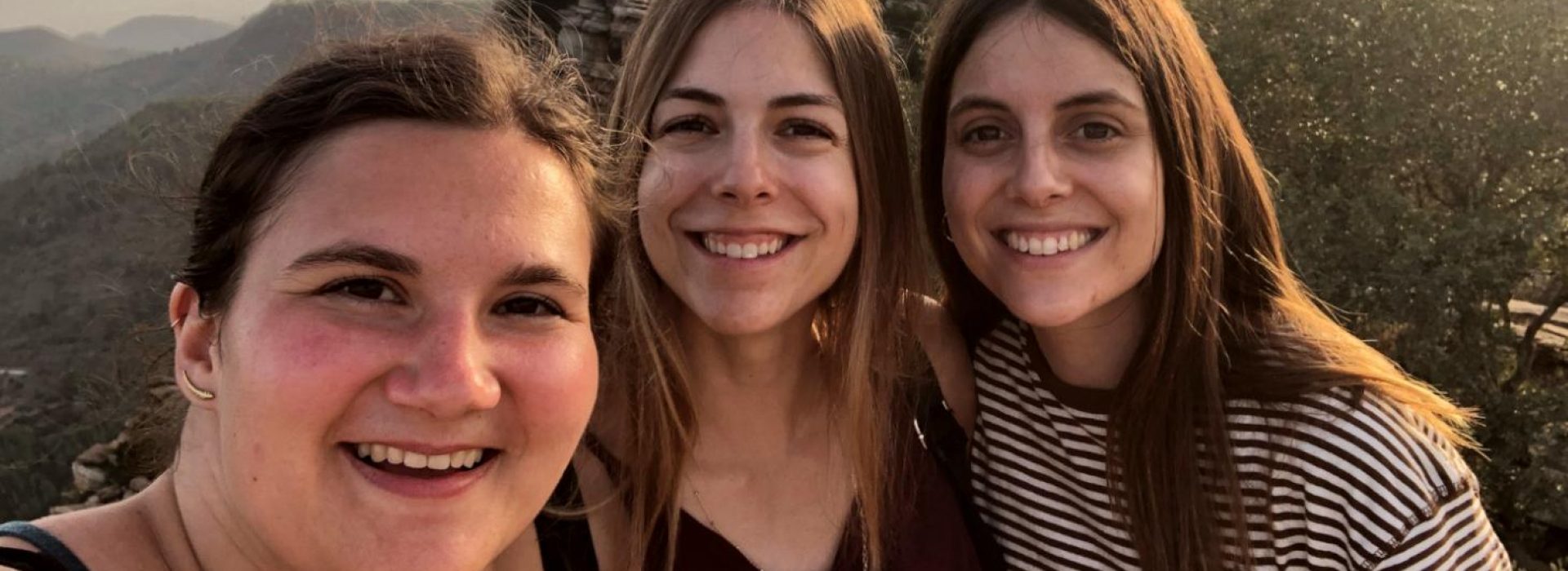 Drei junge Frauen, die im Freien lächeln, im Hintergrund sind bei Sonnenuntergang Berge und Bäume zu sehen.