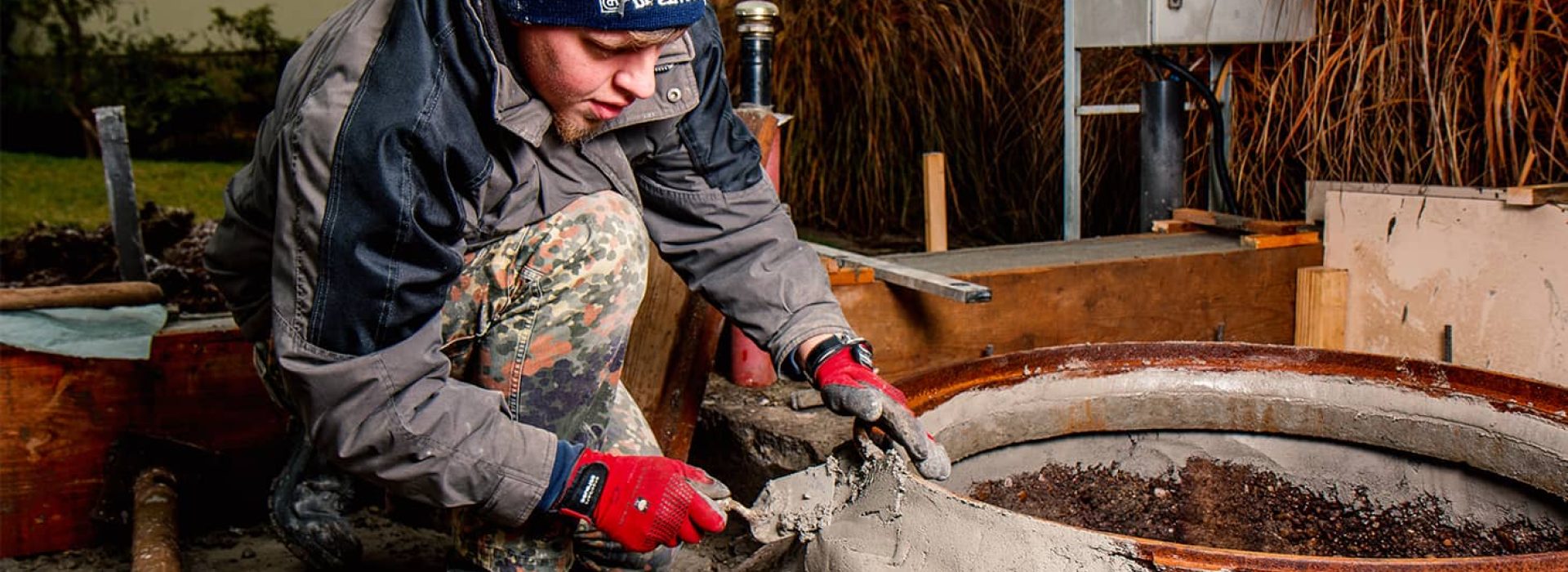 Ein Arbeiter mit Handschuhen und einer Mütze glättet im Freien nassen Beton um eine runde Metallschalung herum.