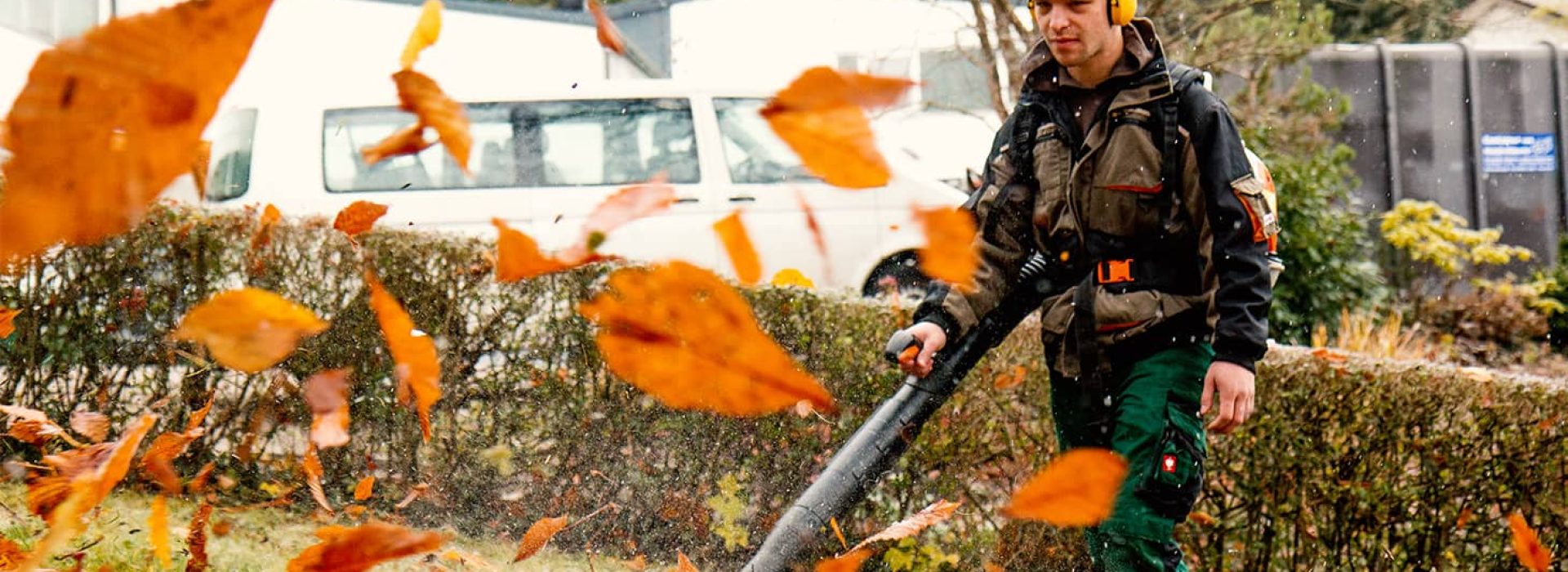 Eine Person räumt mit einem Laubbläser orangefarbene Blätter von einem Rasen und trägt dabei einen Gehörschutz.