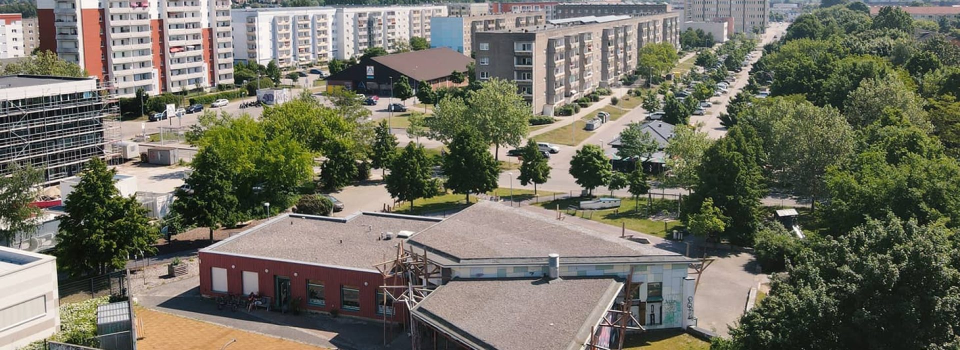Luftaufnahme eines Wohngebiets mit Mehrfamilienhäusern, grünen Bäumen und einigen niedrigen Gebäuden im Vordergrund.