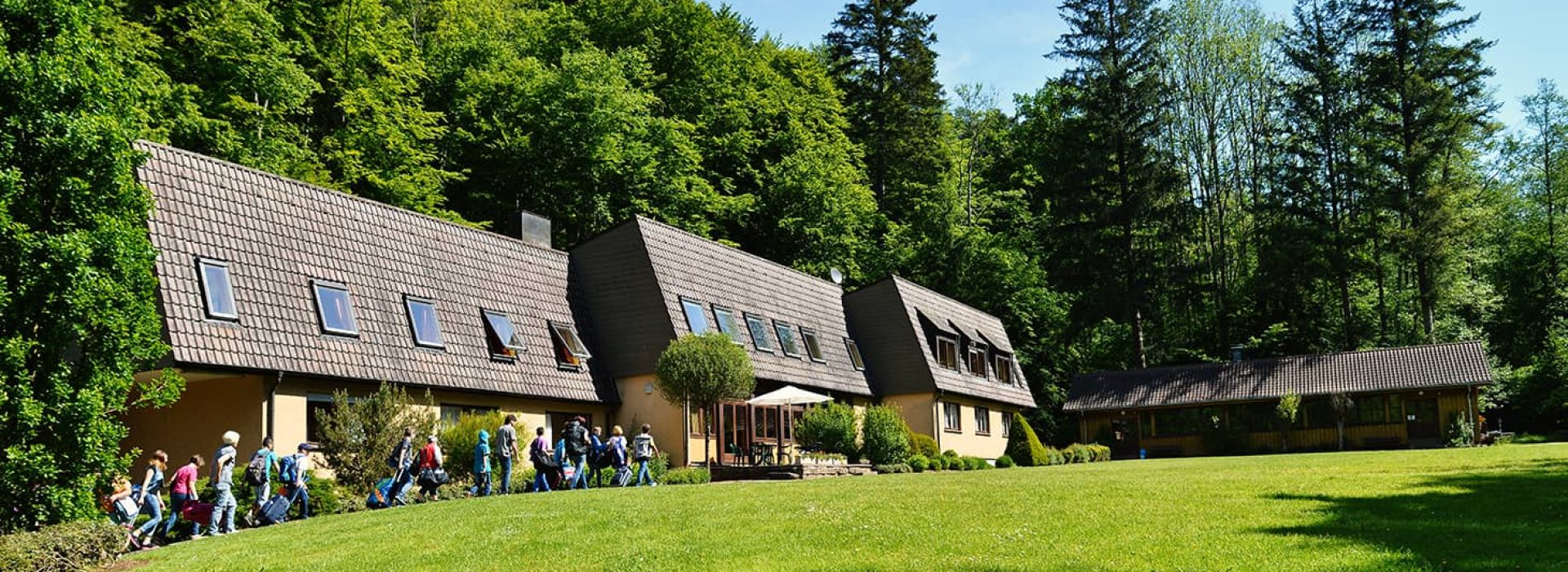 Eine Gruppe spaziert an einem sonnigen Tag über eine Rasenfläche in der Nähe eines großen Gebäudes mit schrägem Dach, das von Bäumen umgeben ist.