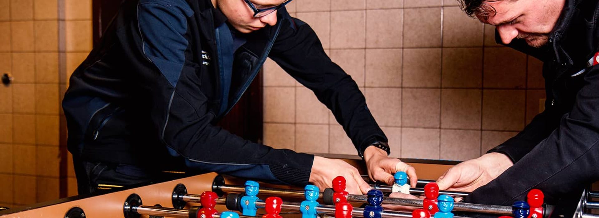 Zwei Personen spielen konzentriert Tischfußball und sind ganz auf ihr Spiel vertieft; sie befinden sich in einem Innenraum mit hellbraunen Fliesenwänden.