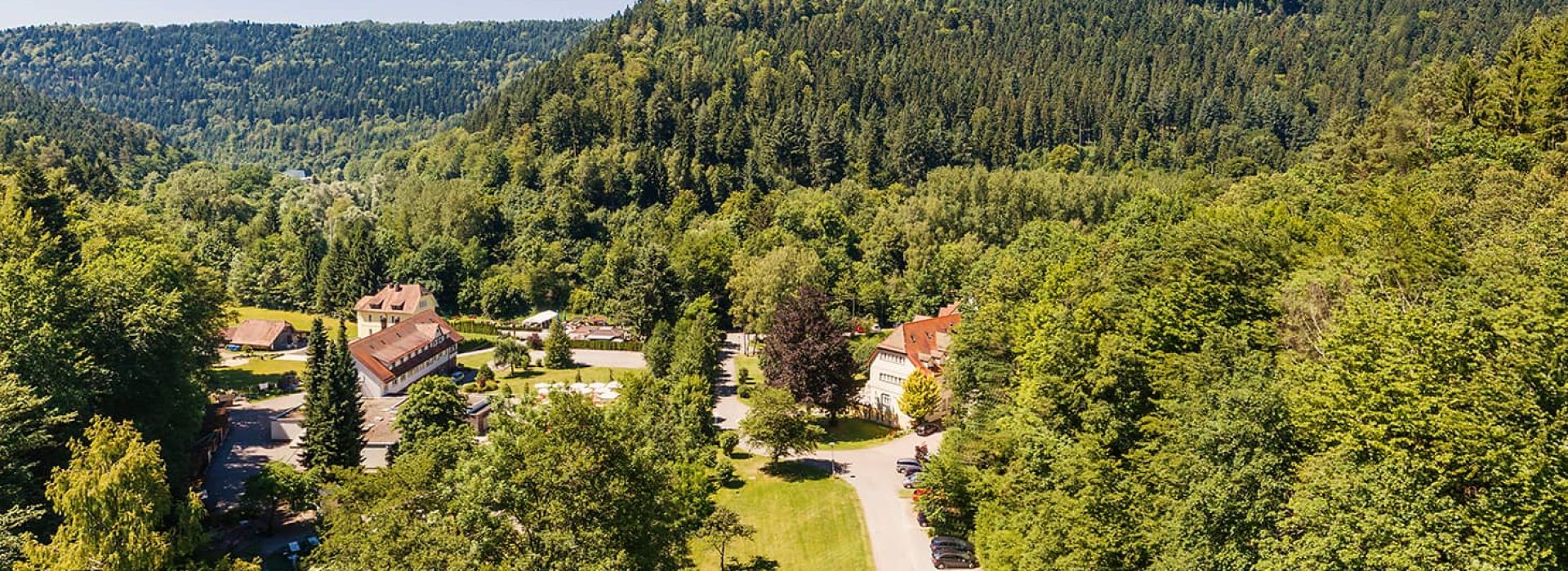 Luftaufnahme eines Dorfes mit Häusern, umgeben von dichtem Wald und grünen Hügeln unter einem strahlend blauen Himmel.
